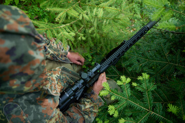 Hunter with rifle wearing camouflage suit sitting in ambush. Camouflaged sniper in the forest in ambush. Military man hide in the woods with rifle.