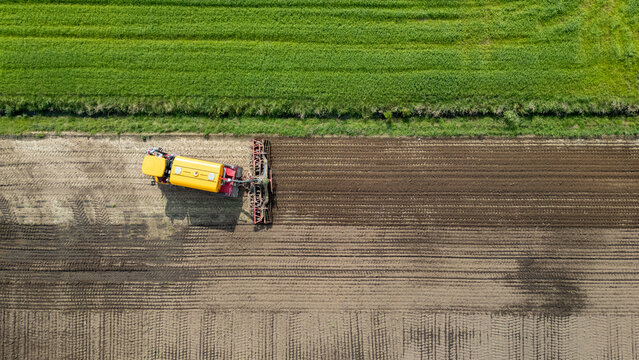 Drone shot of a yellow tractor plowing soil adjacent to lush green fields, ideal for agricultural concepts.