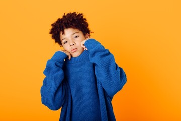 A young african american boy in a blue sweater shyly covers his face with his hands, expressing...