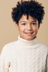 A young african american boy with curly hair wearing a white turtleneck and smiling playfully against a neutral background