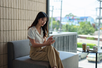 Happy young Asian woman surfing the internet wirelessly on her mobile phone while sitting on the sofa. Enjoy the view in the coffee shop Online shopping through the website.