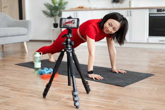 Woman in red workout attire doing push-ups on yoga mat, following online fitness tutorial at home. Exercise equipment like dumbbells and water bottle visible. - Powered by Adobe