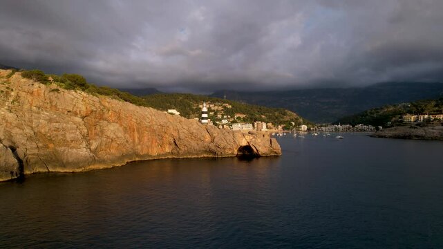 4K Aerial Drone video of popular summer destination, sunset over the cliffs with a lighthouse and a huge port of boats anchored at golden hour with seagulls flying. Port de Soller, Mallorca