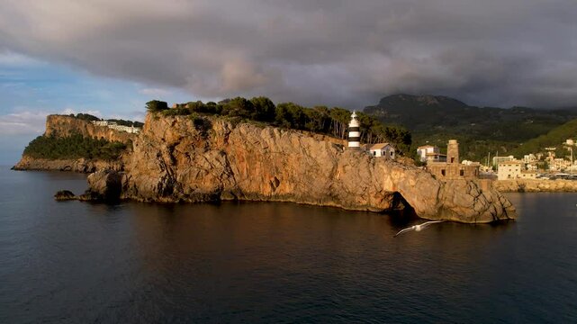 4K Aerial Drone video of popular summer destination, sunset over the cliffs with a lighthouse and a huge port of boats anchored at golden hour with seagulls flying. Port de Soller, Mallorca
