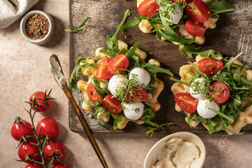 Waffles sandwiches with tomatoes, mozzarella balls, arugula, microgreens on wooden rustic board with ingredients. Homemade breakfast