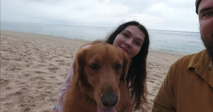 A young woman and a man with a beard take a selfie on an action camera with their Golden Retriever dog. They relax on the beach next to the ocean and record a video for the family. Summer vacation.