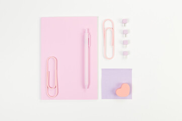 Top view of pink notebook on white background. Sticky note, paperclips, heart, pen and pins. School, office wallpaper. Flat lay, copy space.