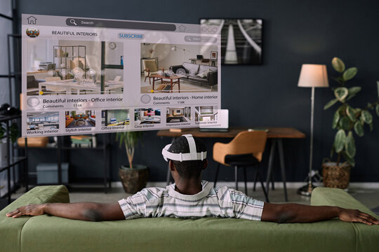 Rear view of unrecognizable Black man wearing VR headset sitting relaxed on sofa in living room surfing Internet - Powered by Adobe