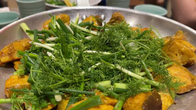 A dish of boneless grilled hemibagrus fish, a type of river catfish fried called Cha Ca La Vong Hanoi, table of diners with fresh dill and spring onion, eating with cilantro