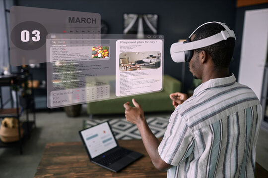 Modern young Black man wearing VR headset standing in living room using time management apps to plan his daily life