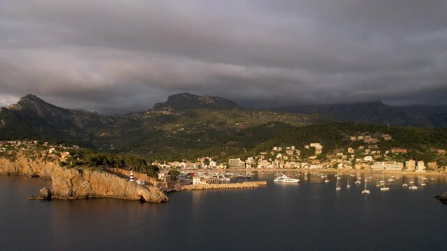 4K Aerial Drone video of popular summer destination, sunset over the cliffs with a lighthouse and a huge port of boats anchored at golden hour with seagulls flying. Port de Soller, Mallorca