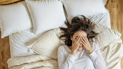 Young woman in her 20s lying in bed with her hands covering her face, looking stressed. She is wearing a white long-sleeve shirt and lying on a bed with multiple pillows
