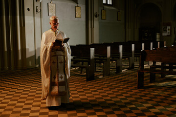 Long shot of senior Catholic priest wearing liturgical vestment standing in empty church building...
