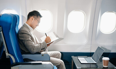 Young Asian executive excels in first class, multitasking with digital tablet, laptop and...