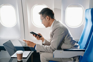 Young Asian executive excels in first class, multitasking with digital tablet, laptop and...