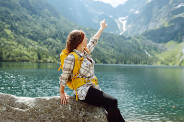 Female tourist with a big yellow backpack enjoys the view of the mountain lake in sunny weather. The concept of hiking, travel, vacation.