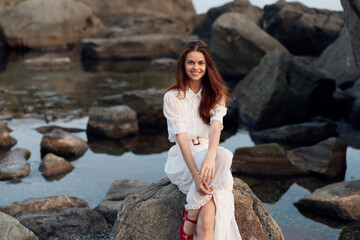 Serene young woman in white dress relaxing on coastal rocks under the summer sun