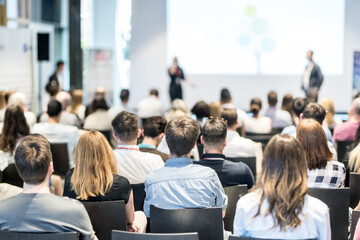 Business and entrepreneurship symposium. Speaker giving a talk at business meeting. Audience in conference hall. Rear view of unrecognized participant in audience.