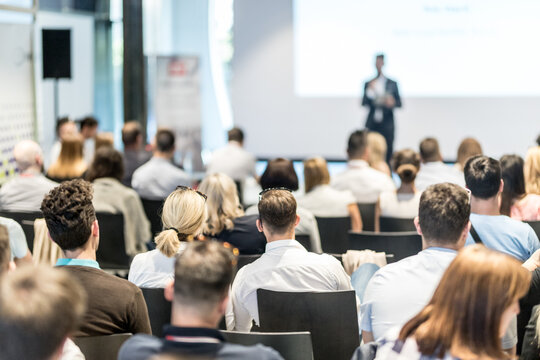 Business and entrepreneurship symposium. Speaker giving a talk at business meeting. Audience in conference hall. Rear view of unrecognized participant in audience.