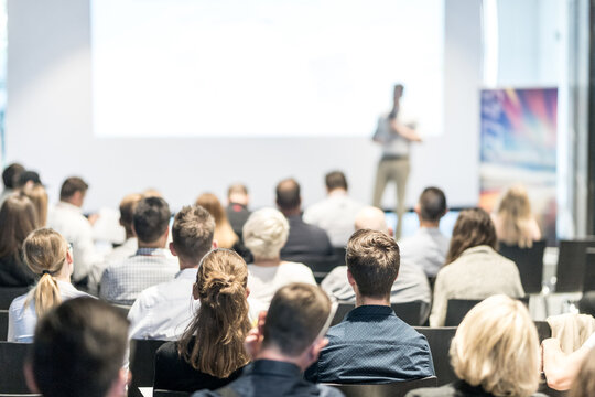 Business and entrepreneurship symposium. Speaker giving a talk at business meeting. Audience in conference hall. Rear view of unrecognized participant in audience.