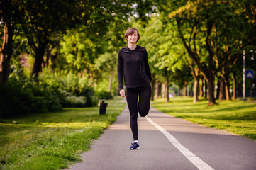 Fototapeta premium A young woman in black sportswear stretching her leg on a park path before running. Highlighting warm-up routines and a healthy outdoor lifestyle.