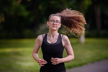A young woman in black sportswear and glasses running in a park, showcasing determination and a...