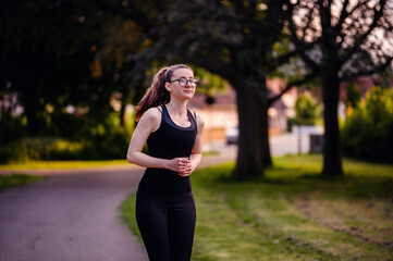 A young woman in black sportswear and glasses jogging on a park path at dusk, showcasing determination and a healthy lifestyle.