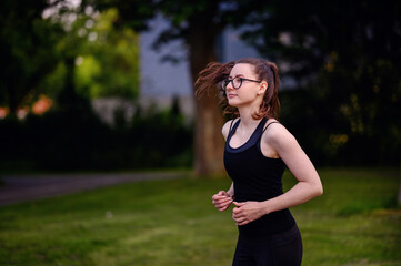 A young woman in black sportswear and glasses jogging in a park, showcasing determination and a healthy lifestyle.