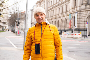 Cheerful middle-aged man, 65 in winter bright yellow puffer jacket and beanie, smiling on urban...