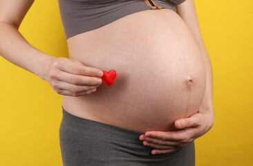 Pregnant woman holding her belly and red heart on yellow background