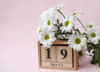 April 19 date on wooden calendar with bouquet of flowers on pink concrete background