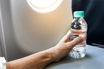 Passenger with dry skin on hand and arm holding a bottle of mineral water next to aircraft cabin window.