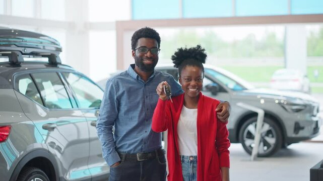 Camera view of African American couple standing in middle of auto salon. Satisfied husband hugging his beautiful wife. Happy lady showing car keys to camera. Man gifting vehicle to his beloved wife.