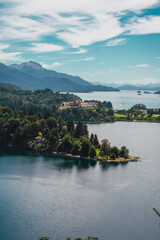 A beautiful lake with a small island in the middle. The island is surrounded by trees and houses. The water is calm and clear. The sky is blue and there are some clouds. The scene is peaceful