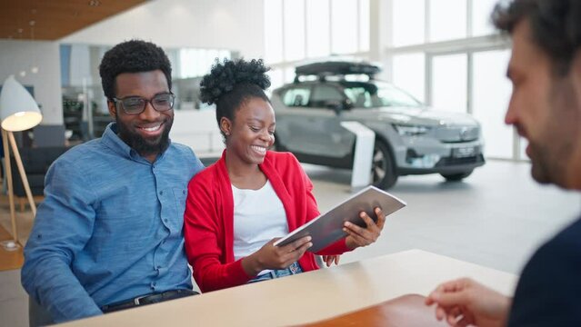 Camera view of African American couple completing agreement with auto dealer for rent or car purchase. Attractive man hugging his wife. Cheerfully smiling and congratulating each other.