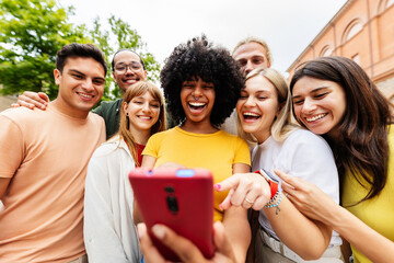 Young group of diverse people laughing using mobile phone at city street. Millennial generation friends having fun watching content in social media app outdoor. Youth and technology lifestyle concept