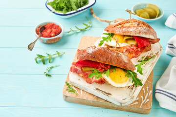 Sandwich layered. English or American freshly prepared picnic sandwich with egg, arugula, bacon, pickles and tomato sause on b turquoise table background on sunny day, copy space