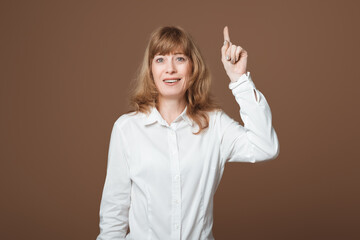 Portrait of attractive Middle-Aged Woman with Blonde Hair Pointing finger at up, showing promo, looking at camera, standing against beige background