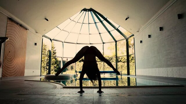 Female gymnast doing handstand and leg movement next to indoor pool, silhouette