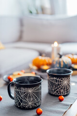 Cozy Halloween plans at home. Hot tea drink in black mugs with spider net pattern, cookies and sweets on the plate, pumpkin decor on the coffee table. Festive autumn, fall mood home decor. Vertical