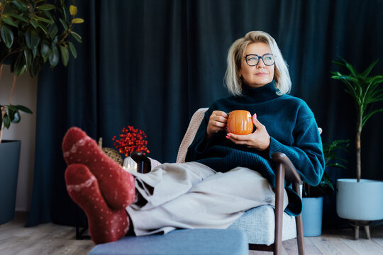 Middle aged woman relaxing with pumpkin shaped cup of hot drink in scandy style hygge interior home with fall mood decor. Lady dreaming, enjoy calm mood without stress, well being alone. Cozy autumn.