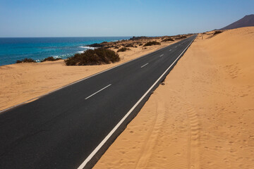 Fototapeta premium Scenic Coastal Highway on Fuerteventura's Golden Shores. Empty Road Cutting Through Desert Sands Alongside Turquoise Atlantic Waters
