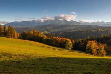 A beautiful autumn landscape of the Tatra Mountains seen from Polish Podhale during the golden hour. The sky is painted in warm hues, with the mountains bathed in golden light.
