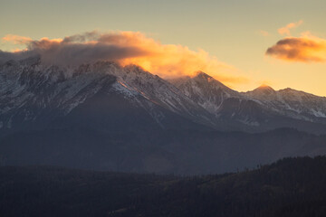 A beautiful autumn landscape of the Tatra Mountains seen from Polish Podhale during the golden hour. The sky is painted in warm hues, with the mountains bathed in golden light.
