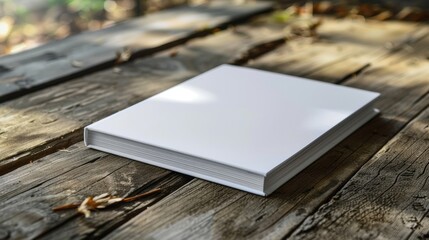 Fototapeta premium A blank white book placed on a rustic wooden table, surrounded by natural light and outdoor elements.