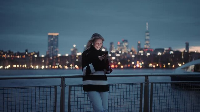 Young blonde woman smiling and texting on her mobile phone by the river in New York at night. Light reflections, boats and NYC skyline in the background. Female standing on the pier by the Hudson rive