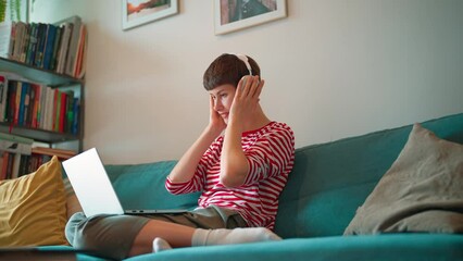 Zooming in on Caucasian female. Wearing new white headphones. Pretty woman using laptop while sitting on soft couch. Searching videos or movie to watch. Getting ready to rest after difficult week.