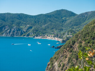 Fototapeta premium Picturesque distant view of Monterosso al Mare with beach, ships and boats seen from Azure Trail to Vernazza with green cliffs, rock, clear blue sky and sea. National park Cinque Terre, Liguria, Italy