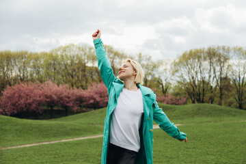 Blond woman in an open turquoise trench coat in a green field.