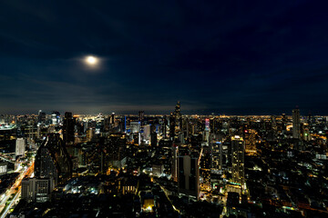 Obraz premium A breathtaking night view of Bangkok's skyline illuminated by city lights and a full moon. The cityscape showcases the blend of modern skyscrapers and vibrant urban life, reflecting the city's dynamic